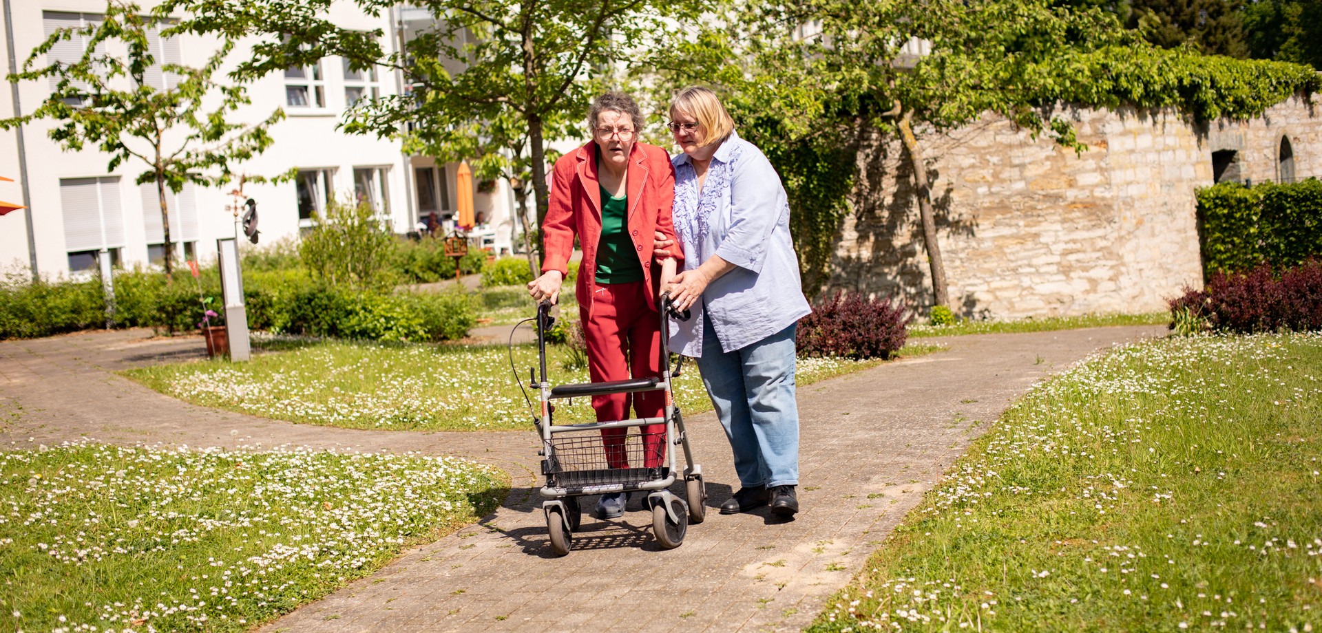 Eine Bewohnerin am Rollator und eine unterstützende Pflegekraft beim Spaziergang im Einrichtungsgarten.